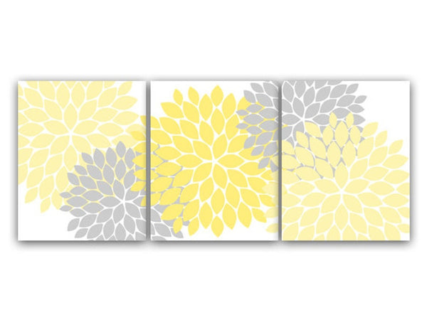 A set of three yellow, light yellow, and gray dahlia floral burst wall art prints, with a white background, designed to be hung on a wall as decor as a triptych. The yellow flower burst is located on the center panel. The left and right panels have a light yellow and a gray flower burst. The flower bursts on the three panels cross over panels. For example, the gray burst on the left panel has petals on the center panel. The same is true for the right panel. The center panel crosses over to the other panels.