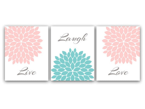 A set of three wall art prints with the words 'Live', 'Laugh', and 'Love' alongside corresponding pink and aqua floral burst designs. The left panel has a  soft pink floral burst filling the upper-right corner and the word "Live" in gray script at the bottom. The center panel has a the word "Laugh" in gray script at the top, and a large teal floral burst filling the bottom half. The right panel has a soft pink floral burst filling the upper-left corner and the word "Love" in gray script at the bottom.